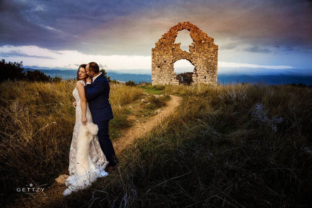 A couple in wedding attire embraces near ancient stone arch ruins on a hilltop at sunset.
