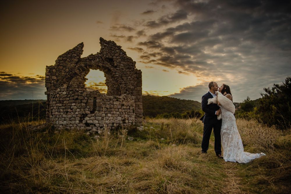 A couple in wedding attire stands near ancient stone ruins during a dramatic golden sunset.