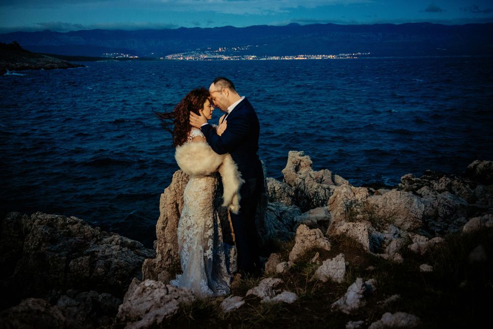 A couple embraces on rocky coastal cliffs during blue hour with city lights twinkling in the background across the water.