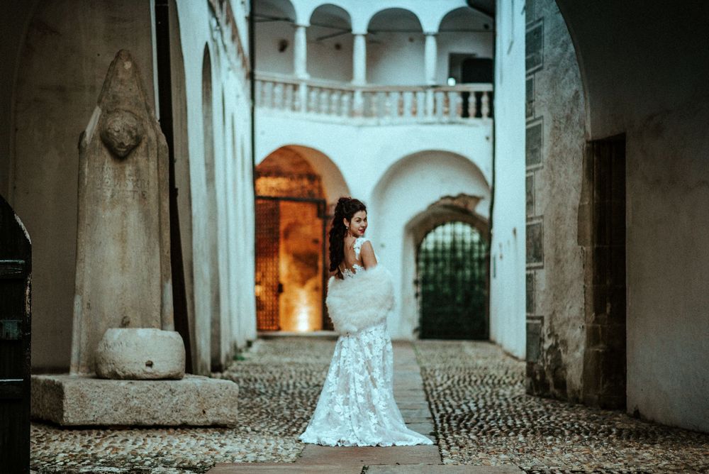 A person in a lace wedding gown poses in an arched courtyard with romantic Mediterranean architecture at dusk.