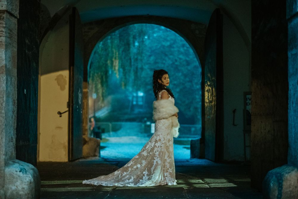 Figure in an elegant lace wedding gown stands beneath a stone archway in a mystical blue-lit setting.