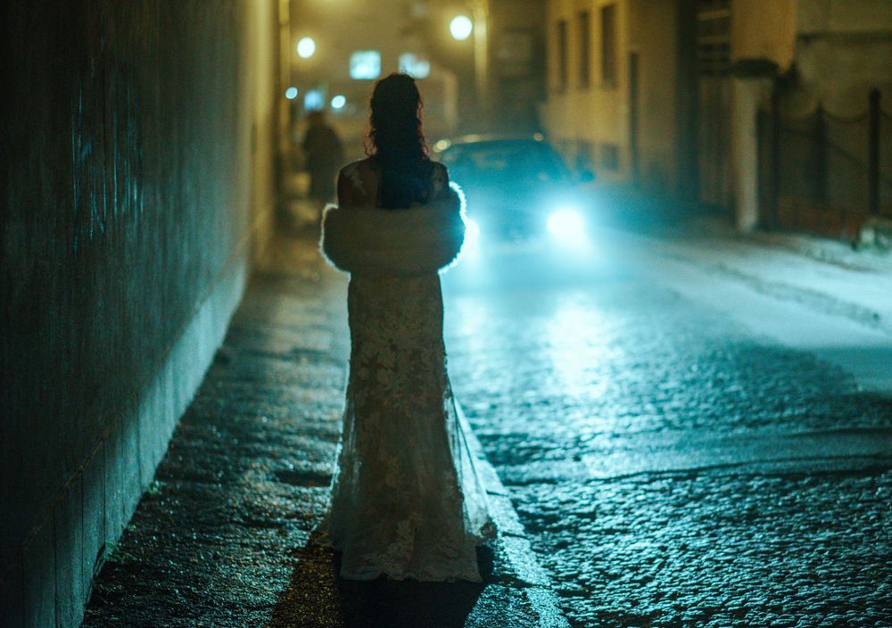 A person in a winter coat stands on a wet city street at night as car headlights approach in the background.