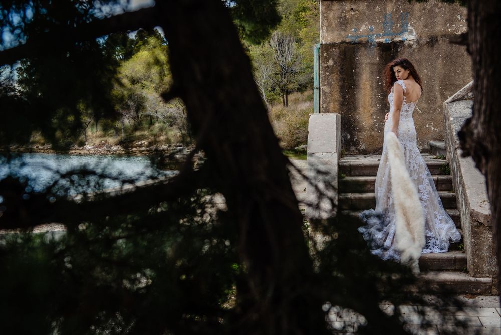 A bride in a white lace wedding dress stands in a stone archway with sunlit trees in the background.