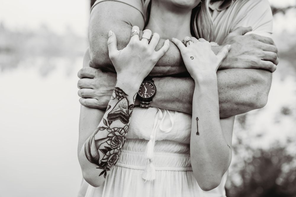 Black and white photo of couple embracing, showing tattooed arms and hands near a lake. Sneads ferry photographer