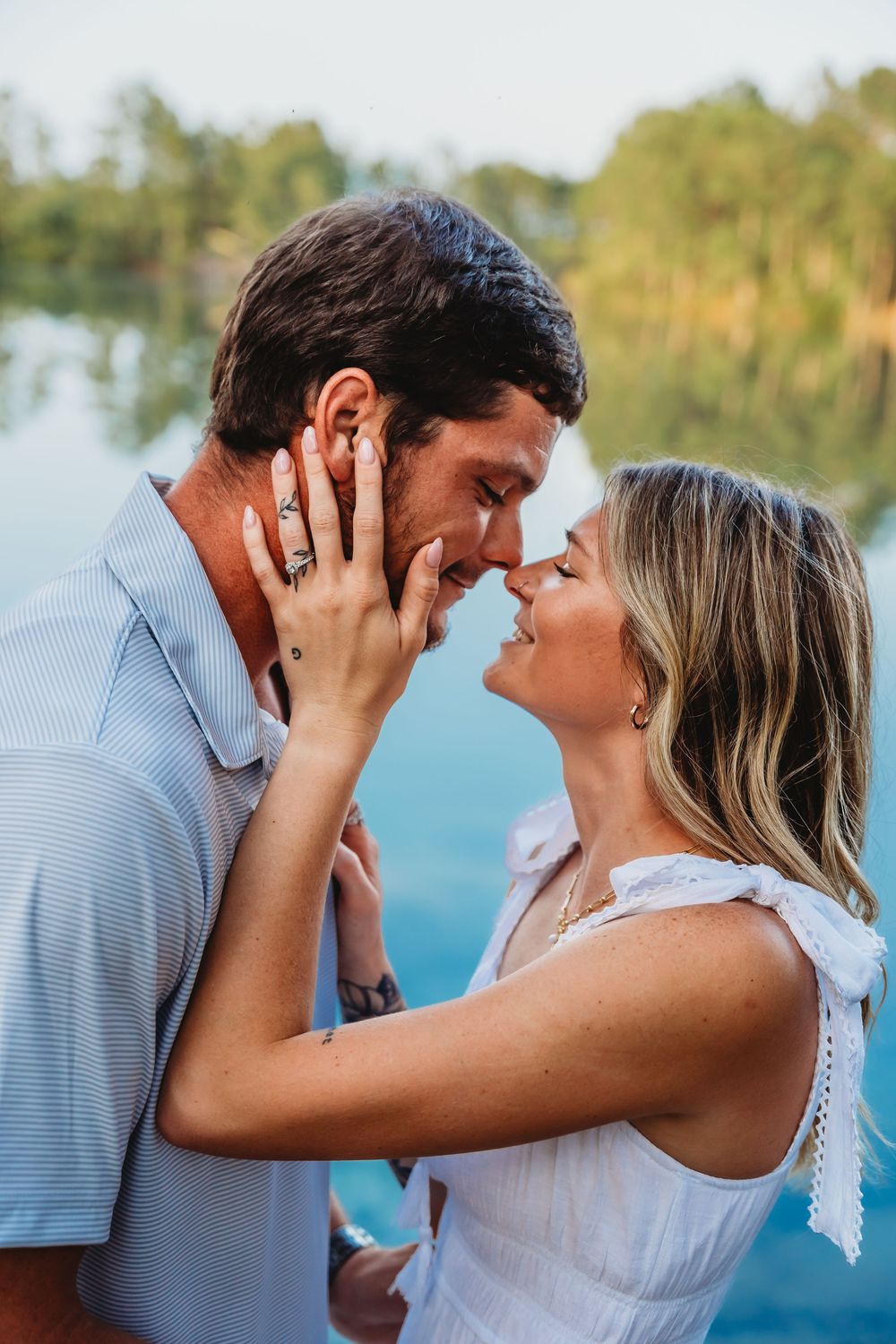 Couple sharing a romantic almost-kiss near a serene blue lake surrounded by lush green trees. Jacksonville, NC