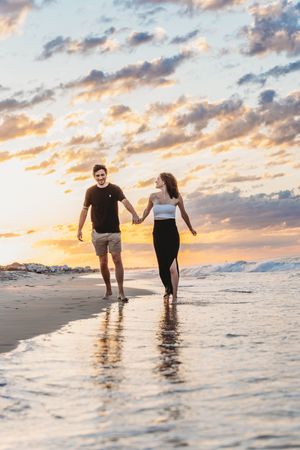 Happy couple holding hands walking along a reflective beach shoreline at golden sunset. Emerald Isle, NC