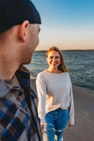 Smiling redhead woman holding hands with man on a beach at sunset with calm water in the background. Jacksonville, NC
