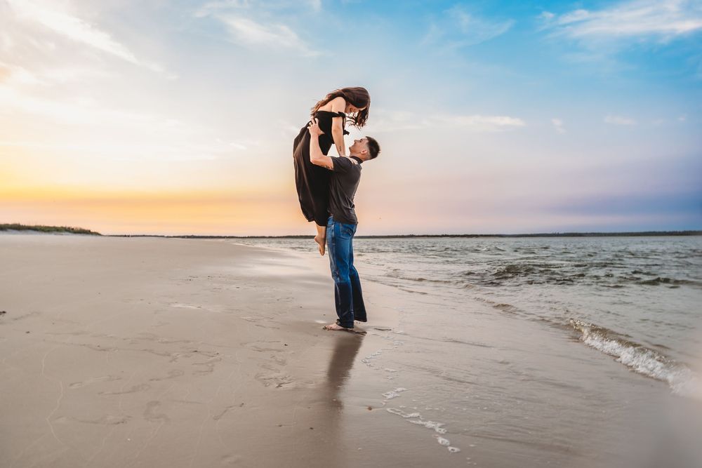Man lifting woman joyfully on a sandy beach during a stunning colorful sunset. Topsail Beach Photographer