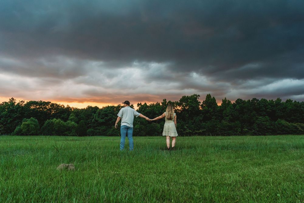 Couple holding hands in a green field under dramatic stormy sunset sky with dark clouds and forest backdrop.