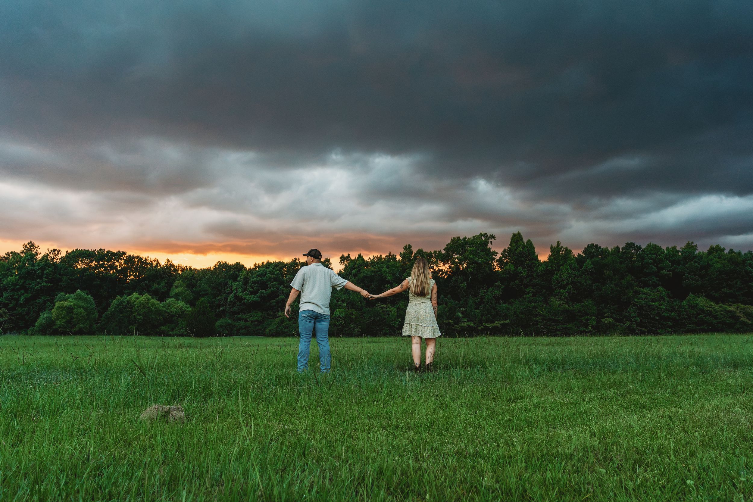 Couple holding hands in a green field under dramatic stormy sunset sky with dark clouds and forest backdrop.