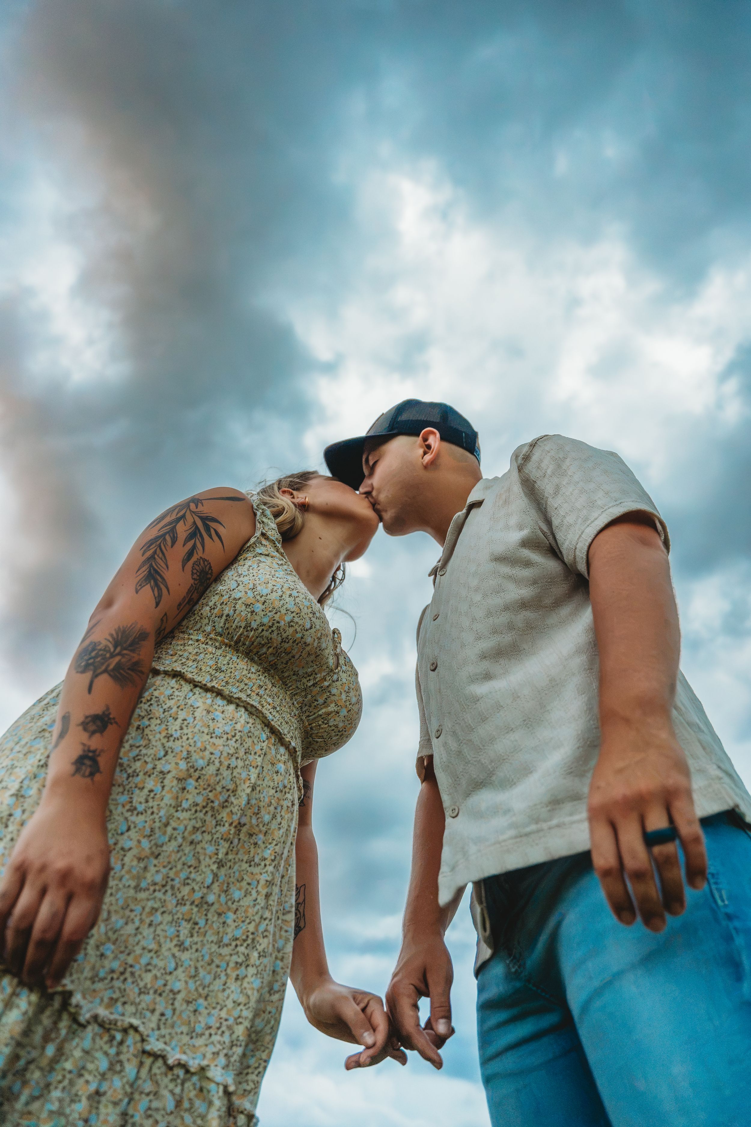 Couple sharing a romantic kiss while holding hands against a dramatic cloudy sky, woman with floral tattoos in sundress.