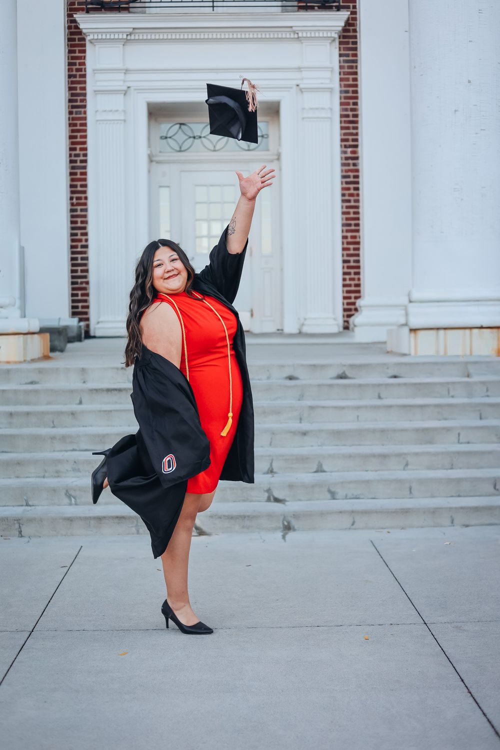 Graduate in red dress and black gown strikes dynamic poses on white stone steps. College graduation photography