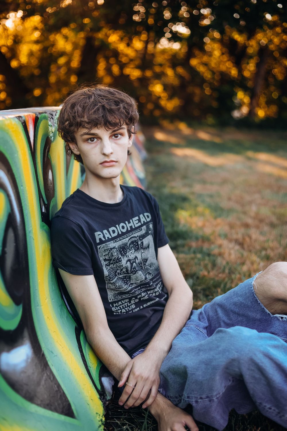A person in a Radiohead t-shirt lounges on a bench in a park during golden hour with autumn foliage in the background.
