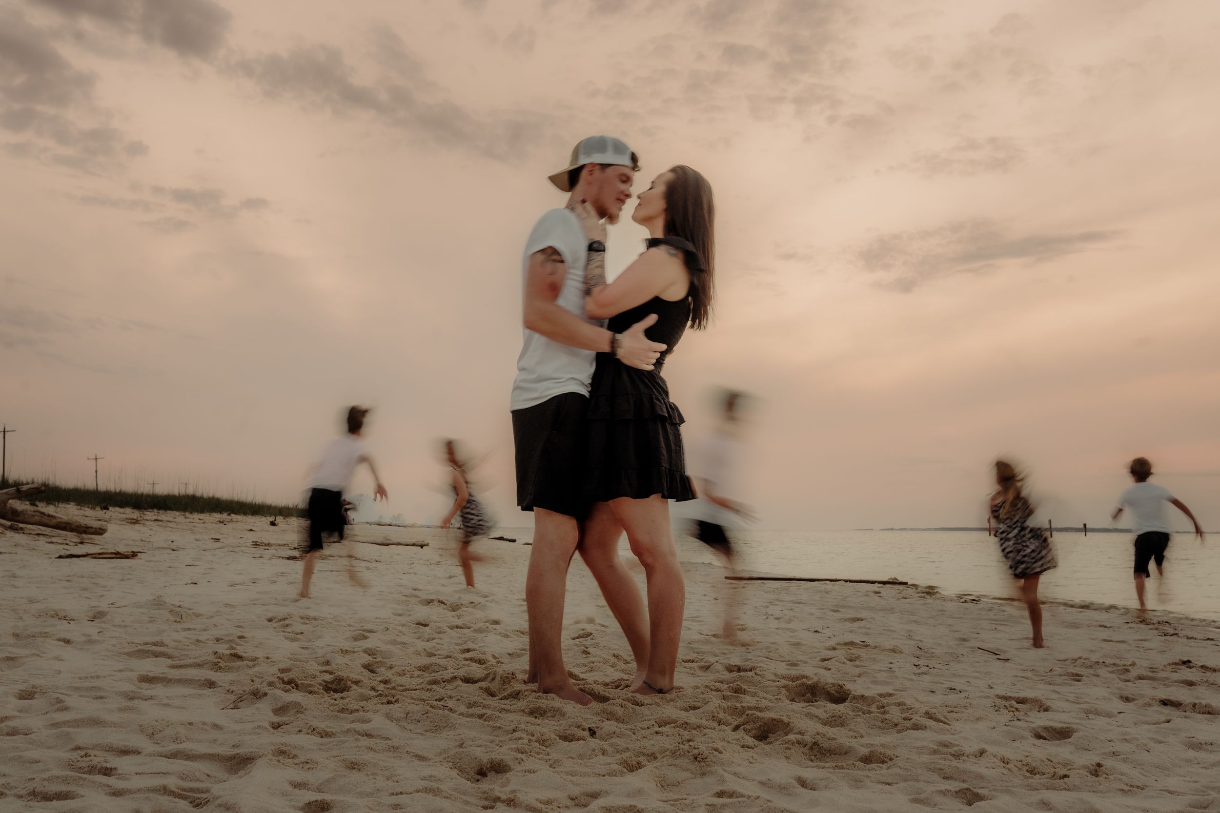 A couple embraces on a beach at dusk while others play in the background.