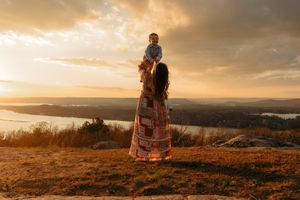 A mother lifts her baby at sunset on a hilltop, wearing a colorful maxi skirt with a scenic river view.