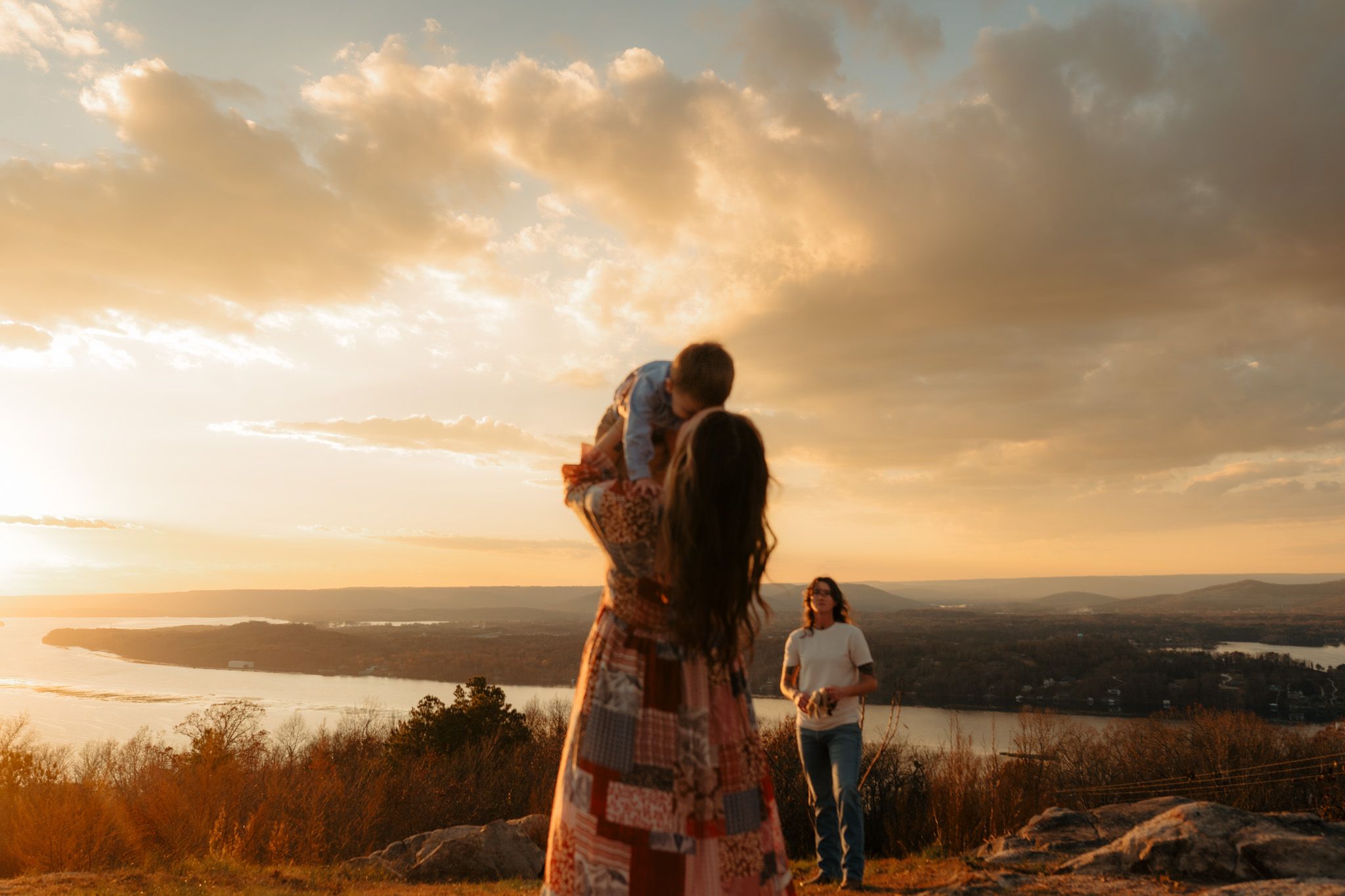 People enjoying a scenic mountain overlook during golden hour sunset with water and landscape views in background.