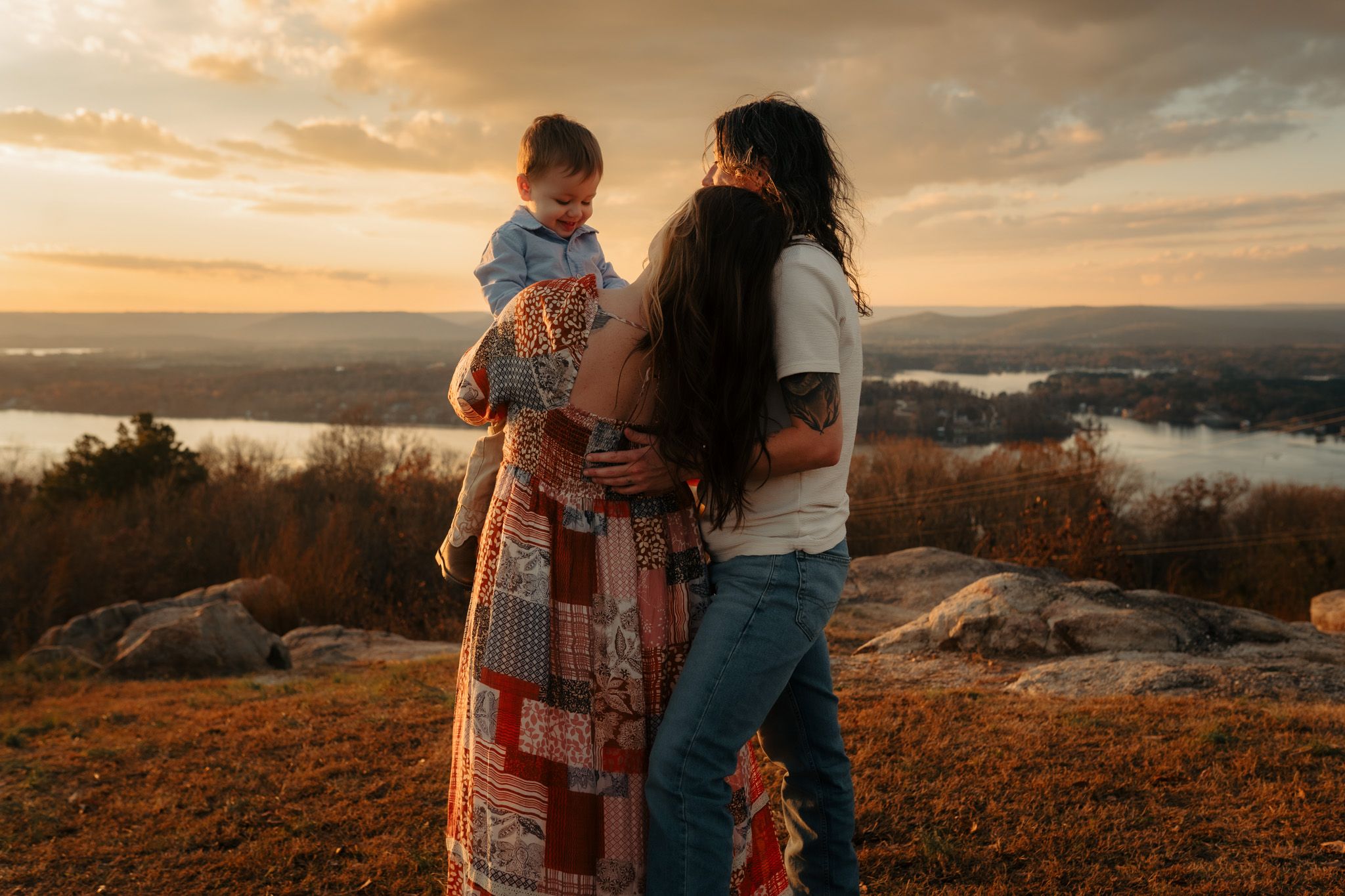 A couple embraces on a hilltop during golden hour sunset with scenic mountain views in the background.