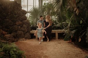 Family relaxing in a lush tropical greenhouse, toddler walking on dirt path while parents sit on a stone bench.