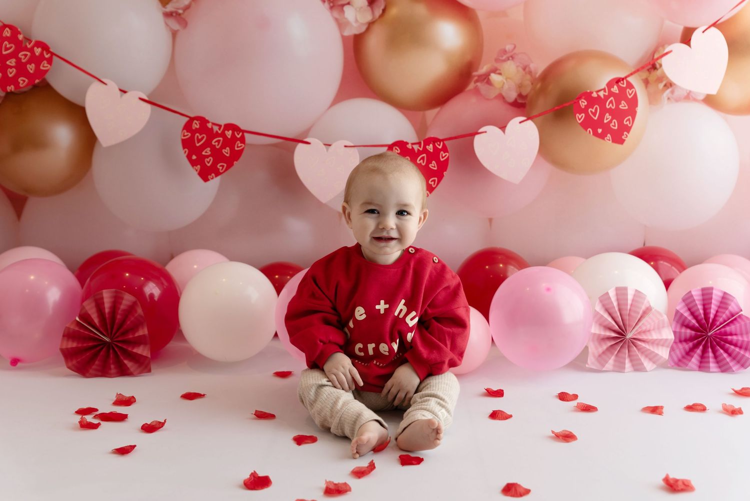 Valentine's day photoshoot setup with red and gold balloons, heart garland, and pink paper decorations.