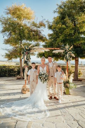 A wedding party stands under a rustic wooden arch with floral arrangements on a stone patio during a sunny day.