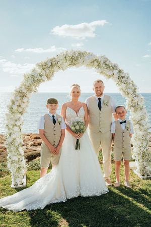 A beachside wedding ceremony setup with a white floral arch and wedding party in beige attire against an ocean backdrop.