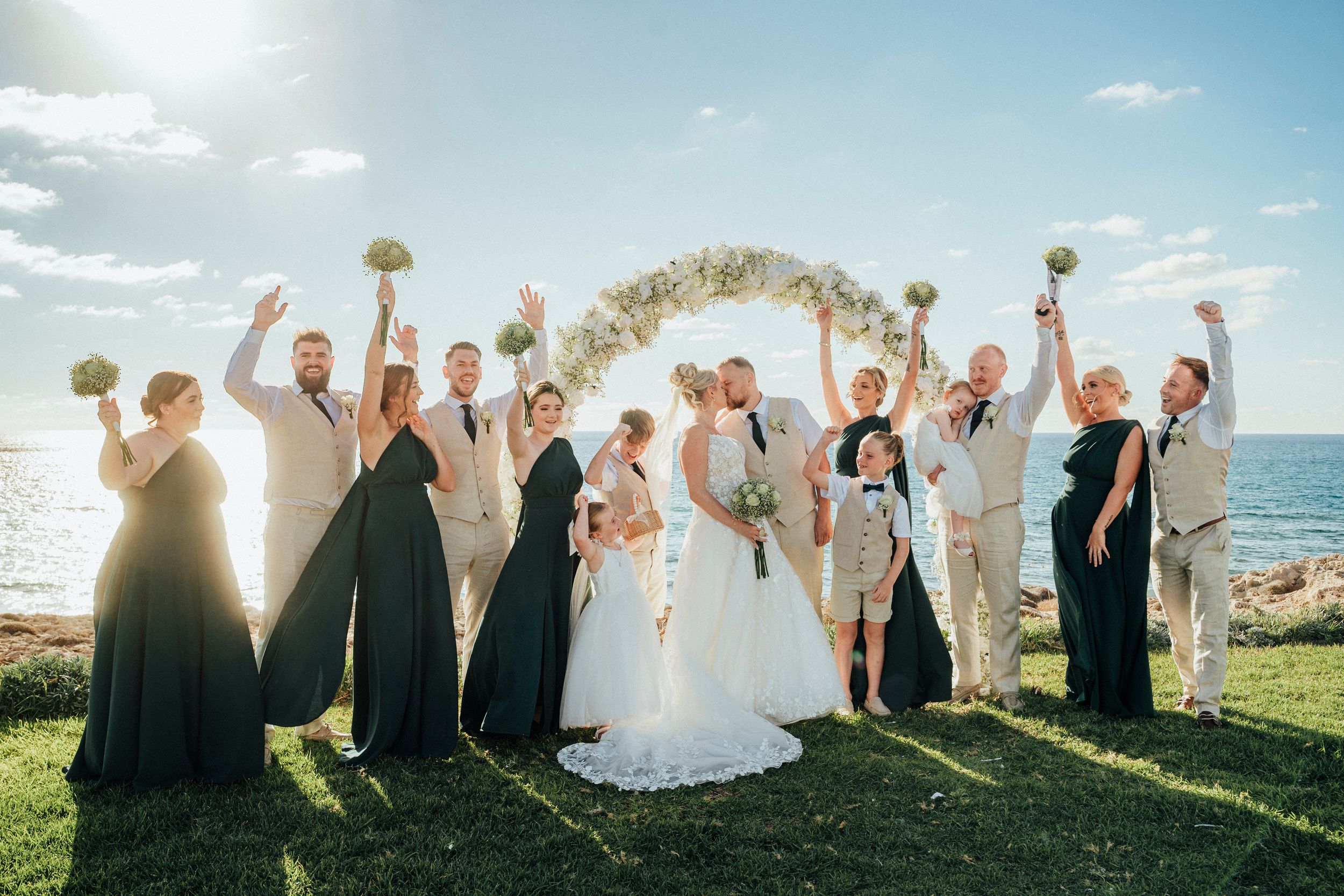 Wedding party stands in a line along the beach with raised arms celebrating during golden hour sunset.