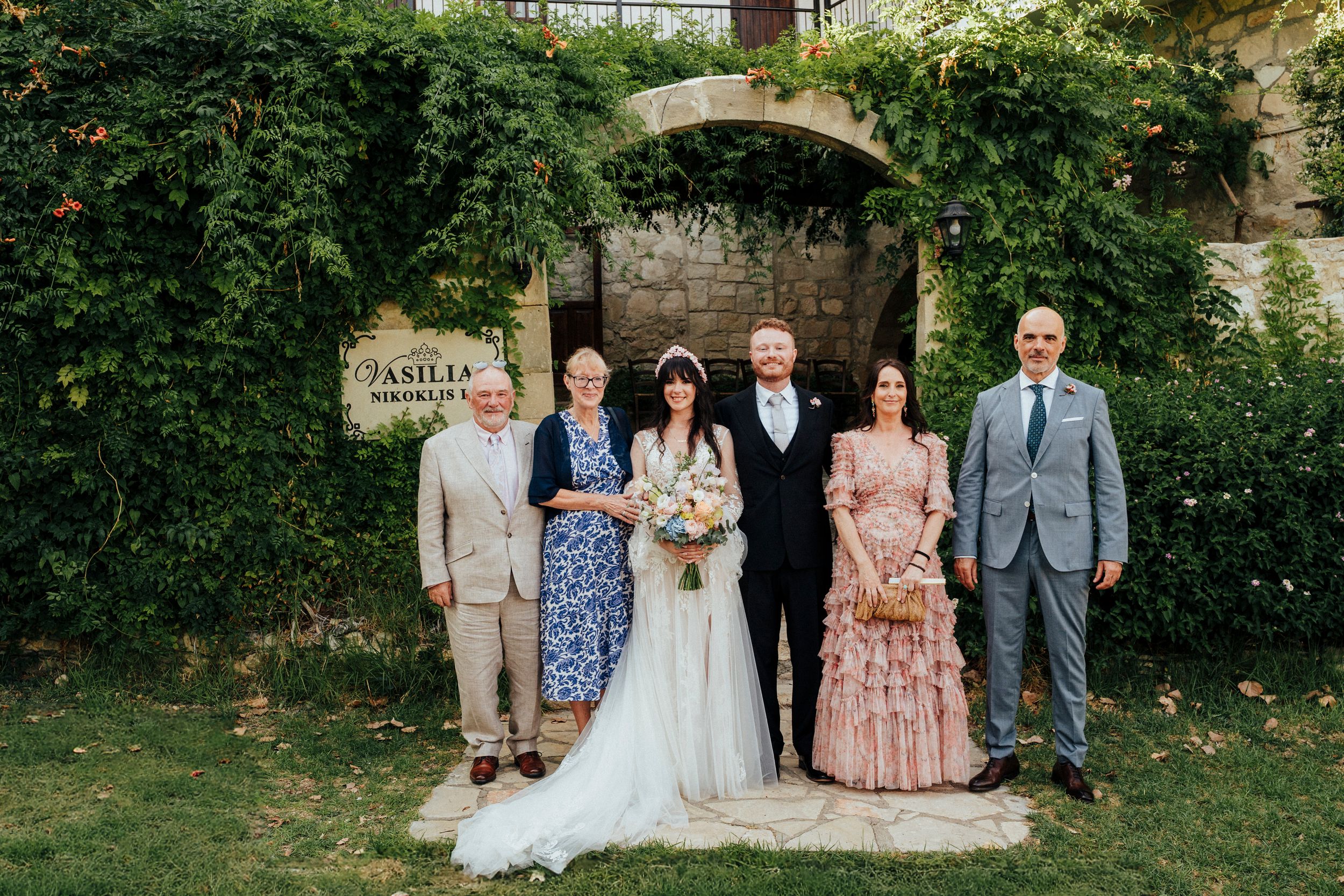 A wedding party poses together in a garden setting with lush greenery and stone walls in the background.