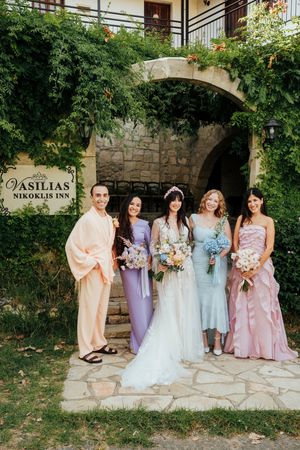 Bridesmaids in pastel colored dresses pose together against stone wall at outdoor wedding venue.