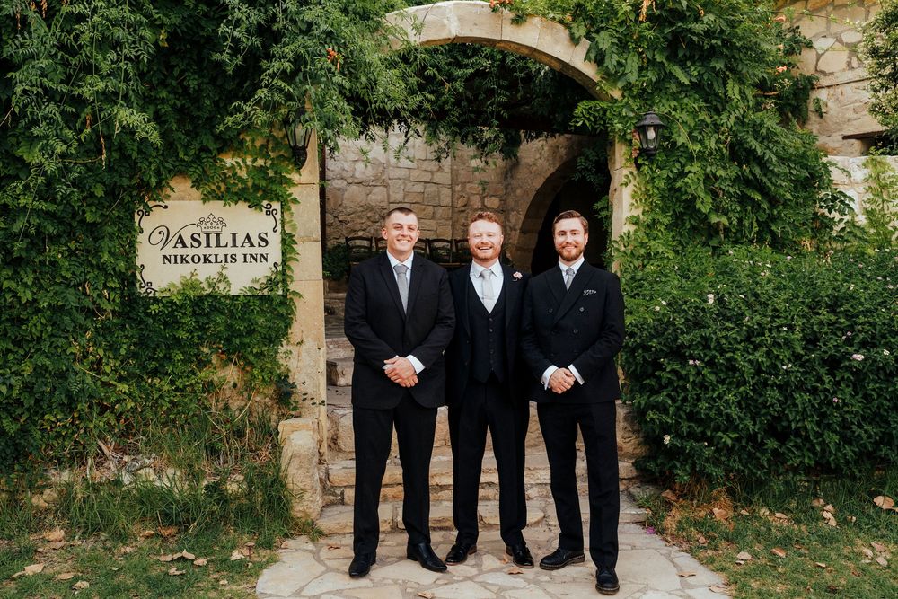 Three men in black suits and ties stand together in front of a stone archway covered with green vines.