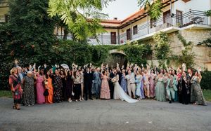Large wedding party gathered outside a white building with greenery, celebrating with raised hands and formal attire.