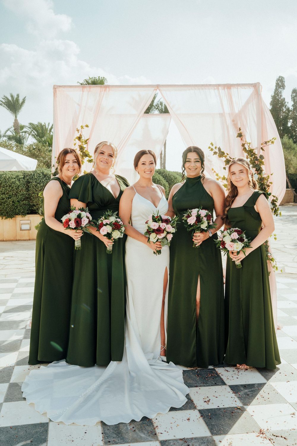 A wedding party in emerald green bridesmaid dresses and white bridal gown pose in front of a pink floral arch outdoors.