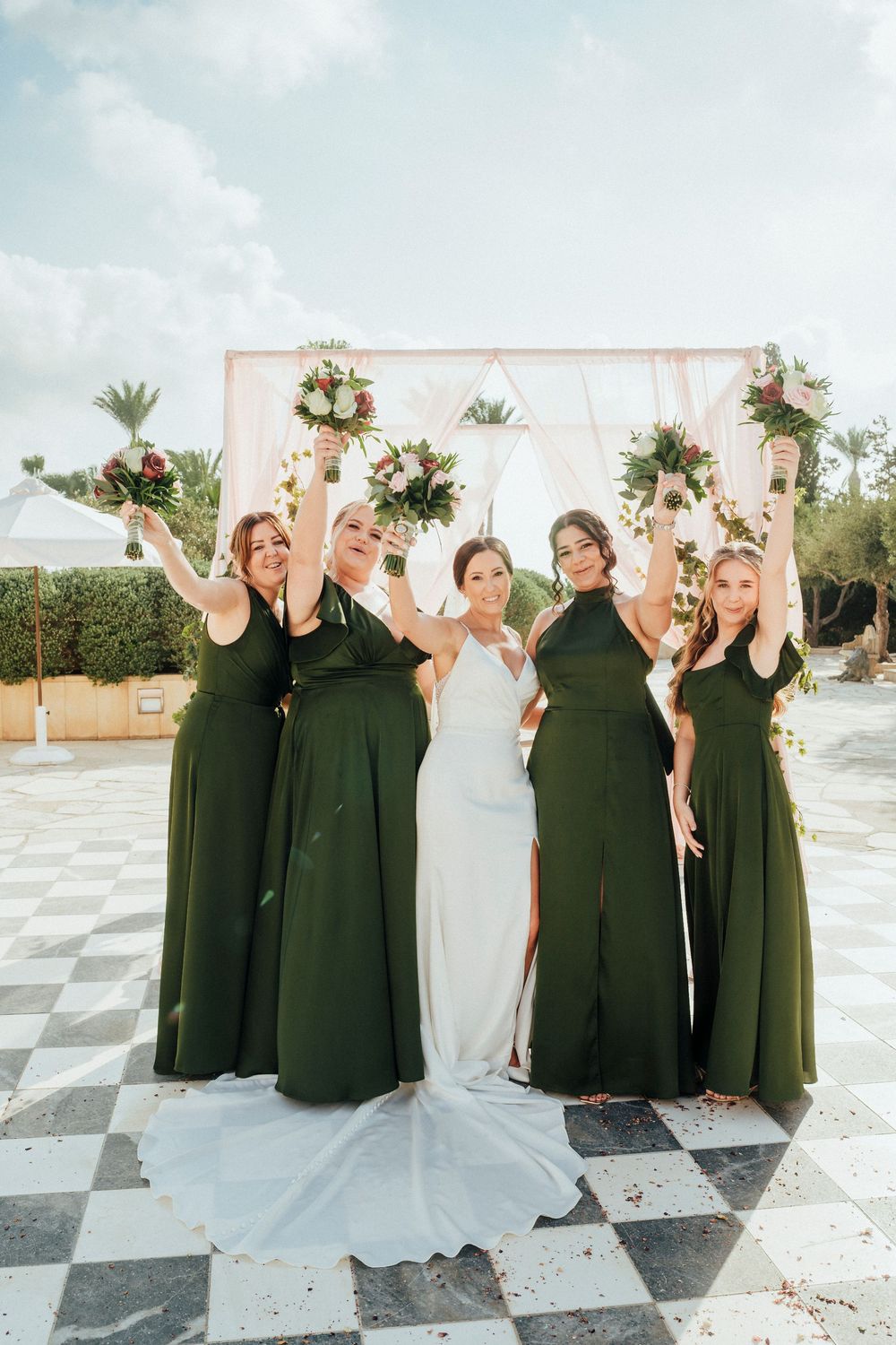 Wedding party poses joyfully on checkered outdoor patio with bridesmaids in dark green dresses and bride in white gown.