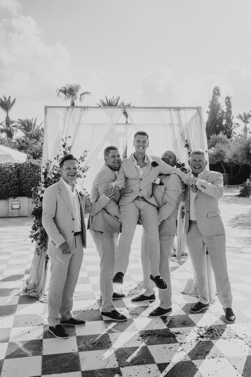 Wedding party in light colored suits strikes playful poses in a sequence of black and white photos outdoors.