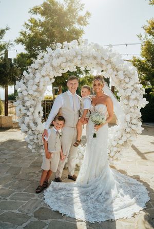 Wedding party poses in front of elegant white floral arch at outdoor venue during sunset.