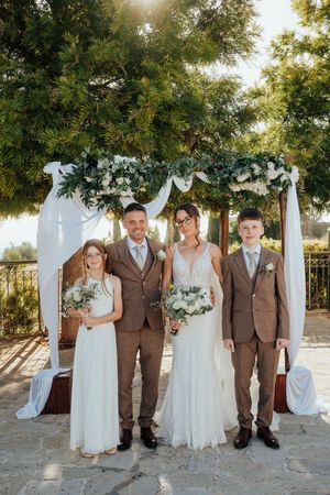 A wedding party in brown and white attire poses together outdoors under trees during a golden sunset.