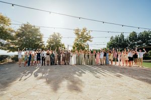 Large group of wedding guests standing together outdoors on a sunny day casting long shadows on the ground.