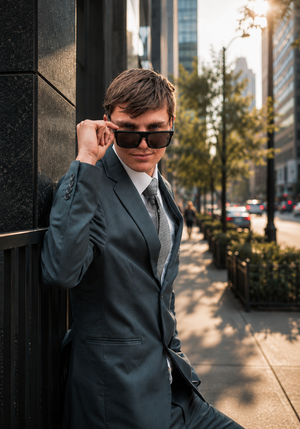 Young man in gray suit and sunglasses leaning against building on urban sidewalk in city.