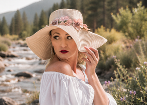 Woman in white off-shoulder top and floral sun hat posing near mountain stream.