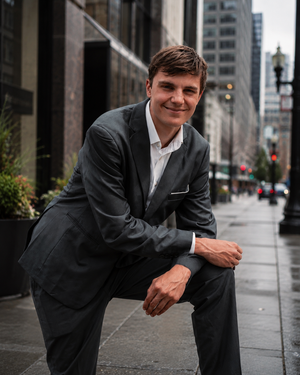 Young man in dark suit smiling while leaning on one leg that is propped up on an urban sidewalk in city setting.