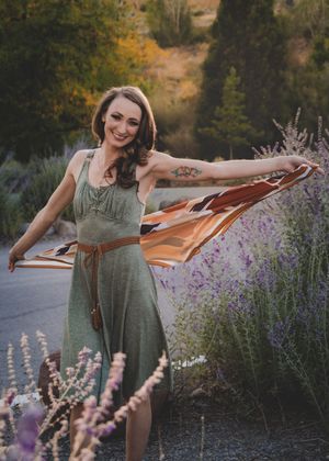 Smiling woman in green dress spreading colorful scarf among lavender flowers outdoors.