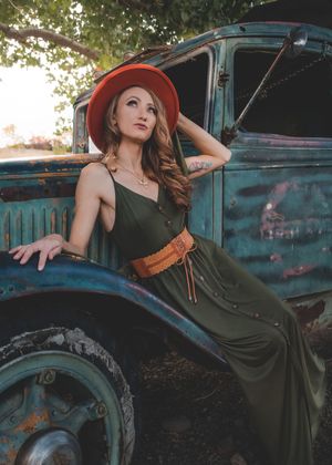 Woman in olive maxi dress and red hat poses against a rustic vintage turquoise truck outdoors.