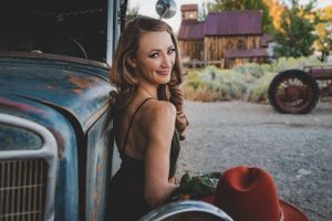 Smiling woman in black dress leans against vintage truck in rural setting.