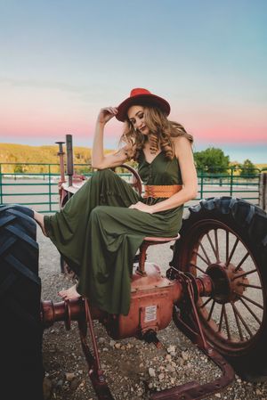 Woman in green dress and red hat poses on vintage red tractor at sunset with hills in background.