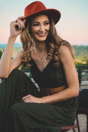 Smiling woman in green jumpsuit and red fedora hat poses outdoors, wearing a dragonfly necklace and brown belt.
