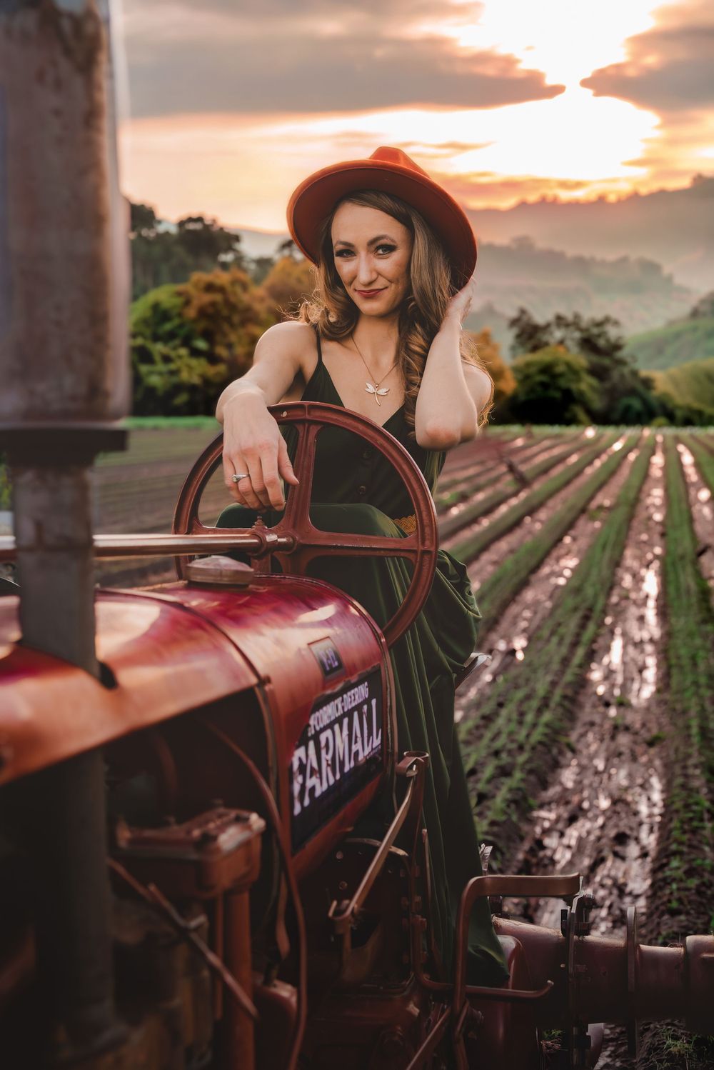 Woman in green dress and hat poses on vintage Farmall tractor at sunset with misty farm fields in background.