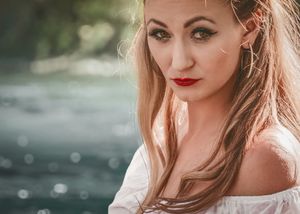 Blonde woman with red lips and green eyes poses outdoors near water in a white off-shoulder top.