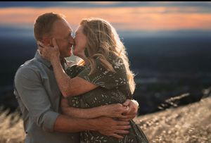 Couple sharing a romantic embrace and kiss at sunset on a hilltop with scenic landscape views.