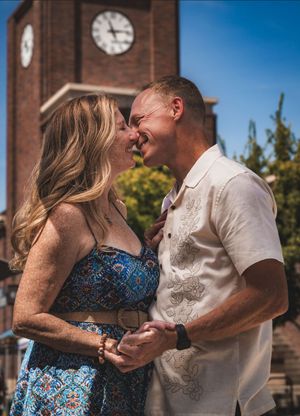 Couple smiling and holding hands in front of a brick clock tower on a sunny day.