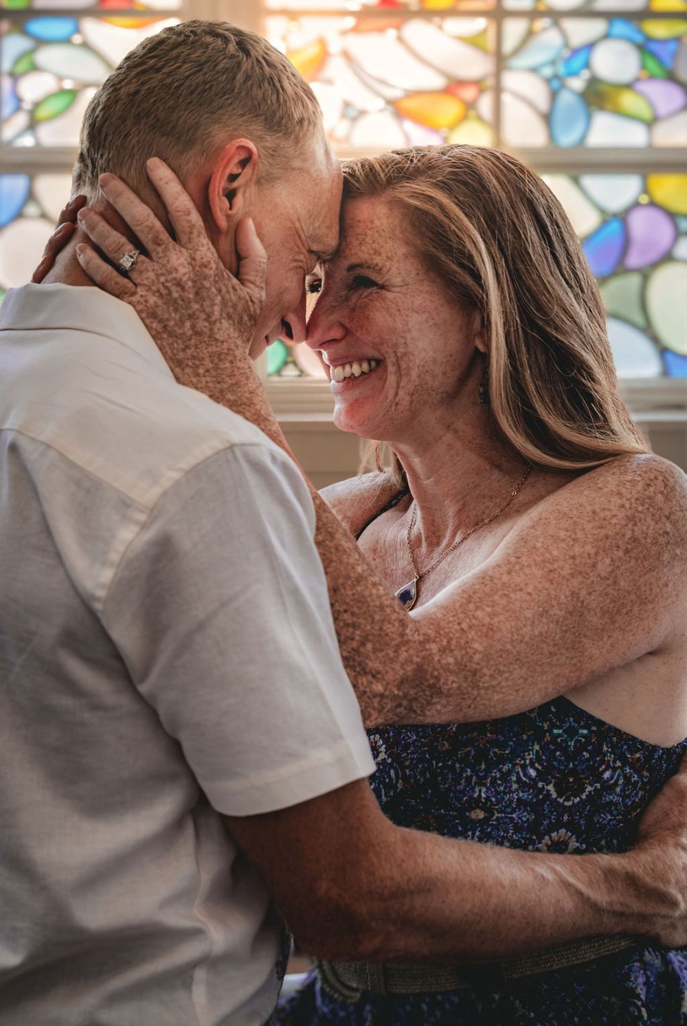 A smiling couple with freckles share a tender forehead-to-forehead moment in front of a colorful stained glass window.