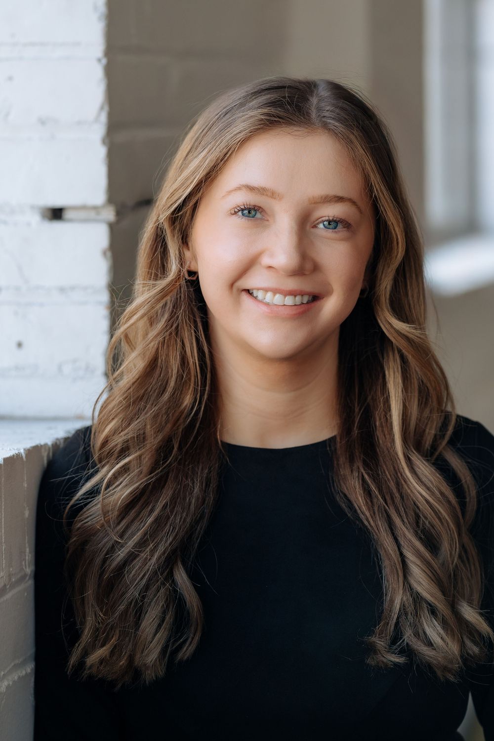 Professional portrait photo of a smiling person with long wavy brown hair wearing a black top against a light wall.